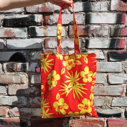Die handgefertigte Tote Tasche hat ein lebendiges Hibiskusblüten-Motiv welches sich über die gesamte Oberfläche der Tasche erstreckt. Die leuchtenden Gelb- und Orangetöne erzeugen einen strahlenden Kontrast und verleihen der Tasche eine fröhliche und sommerliche Ausstrahlung. 
